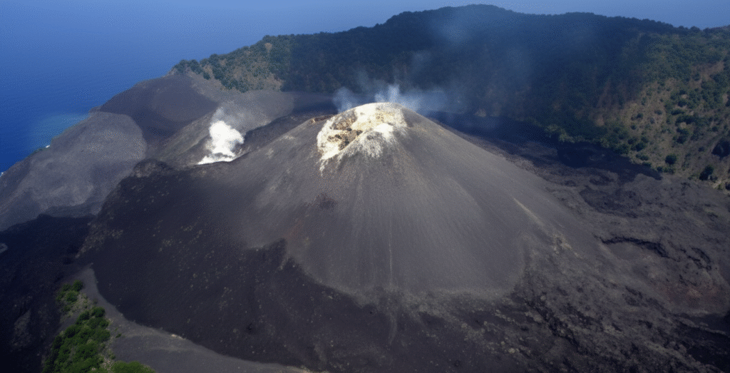 A daytime shot of Barren Island's volcano in the Andaman Sea. A large column of gray smoke and ash rises from the peak against a backdrop of a blue sky with fluffy white clouds. The foreground is dominated by a dark, rugged field of volcanic rock and cooled lava extending to the water. Green, lush vegetation covers the slopes of the surrounding hills to the left and right. The sea is a vibrant blue with small ripples