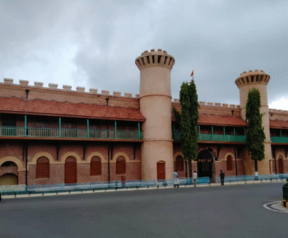 A view of the main entrance to the historic Cellular Jail Andaman, showcasing its red brick facade, two large circular towers, and a red-tiled roof under a cloudy sky.
