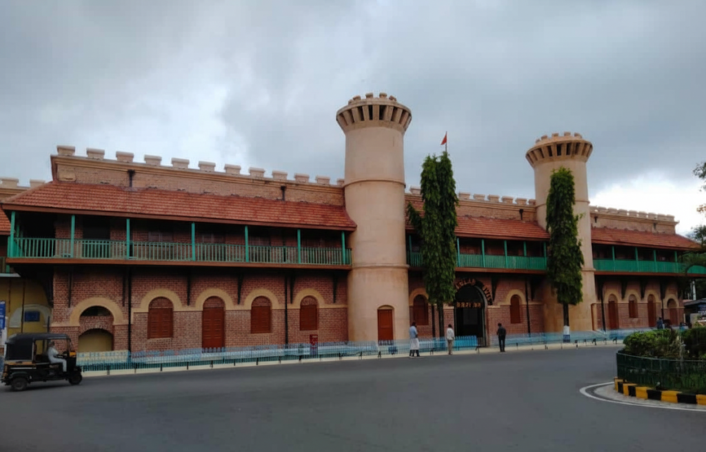 A view of the main entrance to the historic Cellular Jail Andaman, showcasing its red brick facade, two large circular towers, and a red-tiled roof under a cloudy sky.